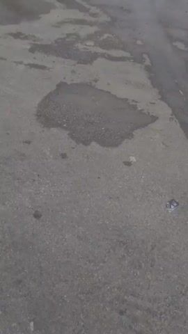 The scene is a gray, damp outdoor area, possibly a parking lot, with several puddles on the ground. The concrete surface is marked with dark, irregular patches, indicating areas of previous water accumulation or stains. In the background, a person dressed in dark clothing and standing next to a white motorcycle with side compartments is visible. The motorcycle is reflected in one of the larger puddles. A second person can be seen at the far left of the frame. The lighting suggests an overcast day.