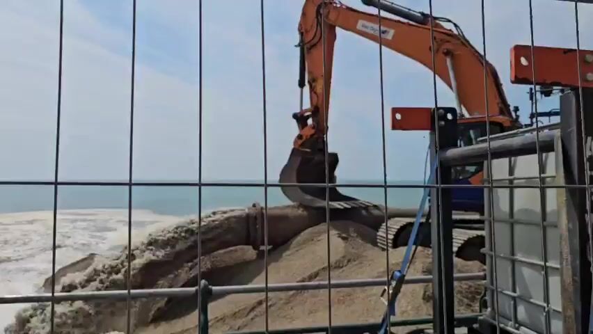 The video shows an excavator with an orange boom and bucket positioned on a sandy area next to the ocean. The excavator's bucket is partially submerged in the water, and a large pipe extends from the excavator into the water, discharging a mixture of sand and water. The water is churning and creating a foamy plume as the sand is deposited. The scene is viewed through a wire fence, with the ocean and a hazy sky in the background. There are no people visible in the frame.