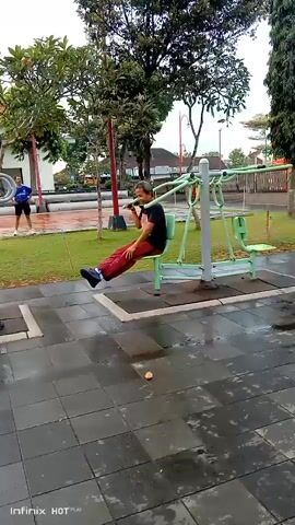 A man sits on an outdoor exercise machine in a park. He is wearing a dark t-shirt and red pants. He is holding a phone to his ear and appears to be talking on it. The exercise machine is green and has silver bars. The ground is paved with dark gray square tiles, and there is a patch of wet grass to the left of the machine. In the background, there are trees, a building, and a street lamp. Two people are walking in the middle ground, and another person is standing in the background to the left. The sky is overcast.