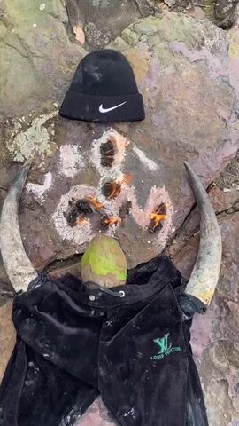This media file displays an outdoor scene on a rocky surface. The arrangement suggests a ritualistic or symbolic setup.

In the center, a flat, reddish-brown rock serves as the primary surface. Scattered on the rock are patches of white powder, with several small fires burning within darker areas, creating an abstract pattern. The flames are orange and cast a warm glow.

Positioned at the top of the rock is a black Nike beanie with the iconic white swoosh logo. Below the fires, two large, curved animal horns are placed symmetrically, pointing outwards from the center of the arrangement. In front of the horns, a green coconut rests. Beneath the coconut and horns, a pair of black velour tracksuit bottoms with a green Louis Vuitton logo on the left leg are spread out, filling the lower part of the frame.

The overall impression is one of a deliberate and symbolic arrangement, possibly a spiritual offering or a form of folk magic. The natural, rocky setting and the items used - the horns, coconut, and clothing - suggest a connection to local traditions or beliefs. The time of day appears to be daytime, with natural light illuminating the scene. There are no explicit indications of weather, but the ground appears dry. The text visible includes the Nike logo on the beanie and the Louis Vuitton logo on the tracksuit bottoms.