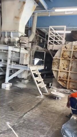 The video shows an industrial setting with two men. One man, wearing a black shirt and brown overalls, is standing on top of some machinery and appears to be using a tool, possibly a grinder, on a large, rusty, circular object. He is wearing a yellow hard hat. The second man, wearing an orange shirt and blue pants, is sitting on the floor to the right of the frame, observing the first man. He is also wearing a blue baseball cap. The environment appears to be a factory or workshop, with various pieces of industrial equipment, pipes, and scaffolding visible. The floor is concrete and dusty.