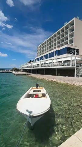 The video shows a small, white boat floating in clear, turquoise water next to a modern, multi-story hotel. The boat is tied to a stone quay and has a small outboard motor and a red fuel can inside. The hotel has a light-colored facade with numerous balconies and a large, open-air dining area along the waterfront. The sky is blue with scattered white clouds. The water is clear enough to see the rocky bottom.