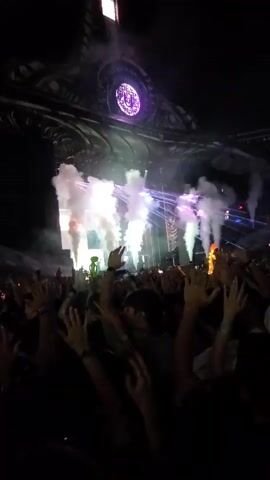 The video shows a crowd of people at a night event, possibly a concert or festival. The stage features elaborate lighting and pyrotechnics, with plumes of smoke and lasers illuminating the darkness. The audience is visible in the foreground, with many hands raised in the air. The overall atmosphere appears to be energetic and celebratory, with a large, complex structure serving as the backdrop for the performance.