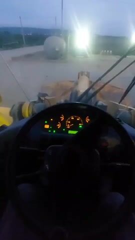 The video begins with a blurry view of a dashboard inside a vehicle. The dashboard has illuminated gauges and indicator lights. The view then shifts to a closer shot of the dashboard, focusing on a hand operating a control lever. The video continues to show the interior of the vehicle with a glimpse of yellow machinery through the window. Finally, the video returns to a wider, blurry view of the vehicle's interior, with bright lights visible in the background, suggesting an outdoor environment at dusk or night. The vehicle appears to be a heavy-duty machine, possibly a loader or tractor, based on the dashboard and the visible external machinery.