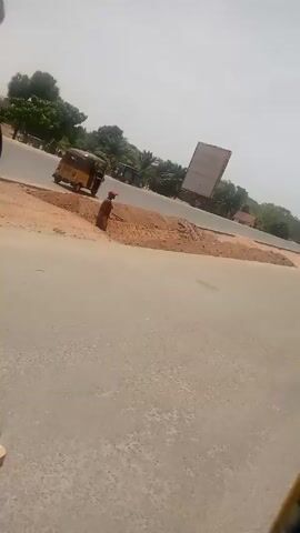The video shows a scene from inside a moving yellow auto rickshaw (tuk-tuk). The camera is positioned to the side, capturing the view of a road junction with other auto rickshaws and motorcycles driving by. There is a pile of dirt at the corner of the intersection and a billboard in the distance. The sky is bright and the scene is generally sunny. A person's leg, clad in jeans, is visible within the auto rickshaw.