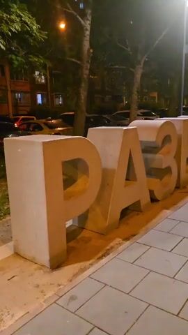 A camera pans right across a row of large, concrete letters that spell out the word "РАЗВИВАЙ" in Russian. The letters are arranged along a paved sidewalk next to a street. Behind the letters, trees and buildings can be seen, along with parked cars. The scene is illuminated by streetlights, indicating it is nighttime.