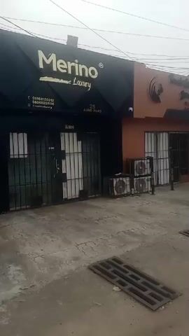 The video shows the exterior of a building with multiple storefronts. The leftmost storefront has a black facade with the "Merino Luxury" logo in gold. Below the logo, there are contact numbers and what appears to be a website address. The entrance to this storefront is covered by metal security bars. Next to it, to the right, is another storefront with an orange facade. This storefront also has a sign with text that is difficult to read due to the image quality, but it appears to include the word "Dream" and "Fashion." In front of this storefront, there are several outdoor air conditioning units stacked together. The area in front of the buildings is paved, and there is a metal grate in the foreground. The sky is overcast. In the distance, on the right side of the video, a few people are visible walking. The overall impression is of a commercial street scene, possibly in a somewhat urban environment.