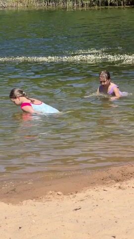 Two young girls are swimming in a lake on a sunny day. They are in shallow water near a sandy shore, with a line of reeds and trees visible in the background. The girl in the foreground is wearing a light blue and pink swimsuit, and the girl behind her is wearing a purple swimsuit. They appear to be playing and enjoying themselves, with one girl looking back at the other. The water is clear and reflects the green of the surrounding vegetation. The sun is high in the sky, suggesting it is midday. The scene is peaceful and idyllic, typical of a summer day spent outdoors in a natural setting.