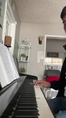 A young man is playing a piano, consulting sheet music displayed on a stand. He is seated on a chair, wearing jeans and a dark jacket. The piano is black, and nearby stands a white decorative shelf with plants. A mirror hangs on the wall with a quote visible in it. The room is illuminated by daylight coming through a window to the left.