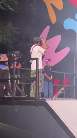 A video shows a skateboarding event. At the beginning, a person in a white t-shirt and camouflage pants stands with their back to the camera, adjusting something on their head. To their right, a young person wearing a purple helmet and a black t-shirt stands holding a drink. A skateboard rests on the platform in front of them. To the far right, another person in a white t-shirt and a white helmet stands with a skateboard. In the background, a large screen displays information about the "WORLD SKATEBOARDING TOUR WORLD CUP ROME 2026". The screen shows a list of standings and a video of a skateboarder. As the video progresses, the camera focuses on the screen which then shows a close-up of a woman named CHENXI CUI, ranked 6th, with her age and stance. Below the screen, on the platform, the three individuals remain. One of the individuals on the platform, wearing a red cap, moves slightly. Another individual, wearing a white t-shirt and a white helmet, also moves. The scene appears to be outdoors at night, with stage lighting illuminating the area.