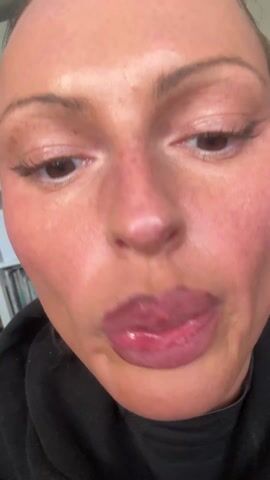 Here is a detailed description of the video. 

The video shows a close-up of a person's face as they apply lip liner. The person has light skin, brown eyes, and dark eyeliner. They are wearing a black sweatshirt. The background is slightly blurred, showing a bookshelf in the distance. The person uses a light pink pencil to line their lips.