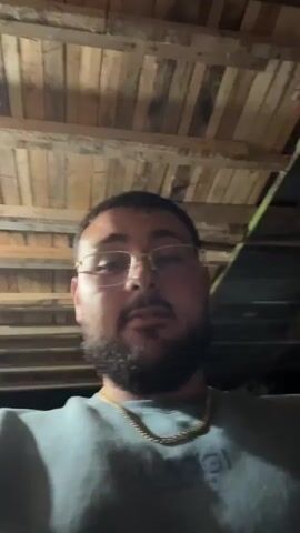 A man with a beard and glasses is looking at the camera. He is wearing a grey t-shirt and a gold chain. The background appears to be a wooden ceiling. The man makes a hand gesture with his right hand.
