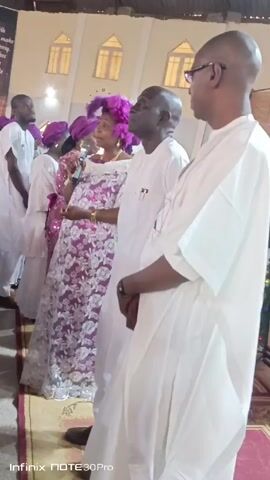 The video shows a group of people standing indoors, likely in a church or large hall. A woman in a purple lace dress with a matching headpiece is speaking into a microphone, while other people, mostly men, stand beside her. They are all dressed in white traditional clothing. The room has several windows in the upper portion of the wall. A sign is visible in the background. The lighting is somewhat dim. The camera is stationary and slightly off-center. At the bottom of the screen, the words "Infinix NOTE 30 Pro" are visible.