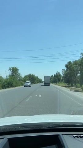 The video shows a view from inside a vehicle driving on a two-lane road on a clear, sunny day. The road is paved and marked with white lines. Trees and greenery line both sides of the road. In the distance, a large white truck is visible in the right lane, moving away from the camera. A silver car is in the left lane, also moving away from the camera. A digital readout displaying "79 km/h" is superimposed on the lower center of the screen, fluctuating slightly throughout the clip. The front dashboard of the vehicle from which the video is recorded is partially visible at the bottom of the frame. The sky is a clear, bright blue with a few faint lines, possibly power lines, visible.