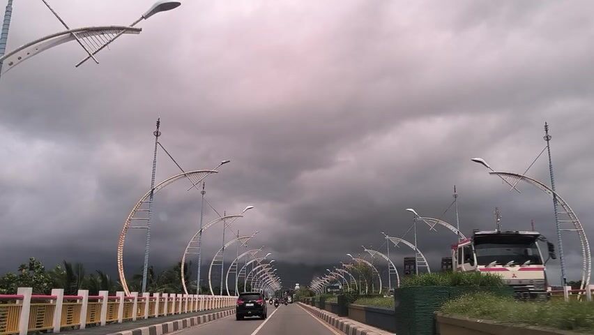 The image shows a view from a moving vehicle on a straight road, possibly a highway or an elevated bridge section, stretching into the distance. The road has at least two lanes in the direction of travel, bordered by yellow and white striped guardrails on both sides.

A prominent feature of the scene is the series of ornate street lights or decorative archways lining both sides of the road. These structures have a distinctive crescent or boat-like design, creating a rhythmic visual pattern along the thoroughfare.

Behind the decorative street furniture, green vegetation, including grass and trees, is visible on either side. Palm trees can be specifically identified on the right. A black car is seen driving away in the far distance on the road, while a blue and white bus is partially visible on the left side. The text "MEDAN" and "BUS" are discernible on the side of the bus.

The sky is entirely overcast with dark, heavy grey clouds, suggesting a gloomy or pre-rainy daytime atmosphere. The perspective indicates that the image was captured while traveling along the road. The street decorations and bus text provide location cues, with "MEDAN" pointing towards a route or destination within Indonesia.