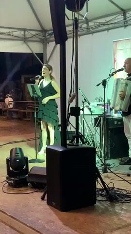 The video shows two musicians performing on a stage under a white tent. A woman on the left is singing into a microphone, holding a sheet of music. She is wearing a dark, tiered dress and sneakers. A man on the right is playing an accordion. He is wearing a dark vest over a white shirt and white pants. Behind them, a soundboard and other audio equipment are visible. A banner with text is hanging on the right side of the stage. The lighting appears to be red and white. The stage floor is visible in the foreground, and beyond the stage, a blurry outdoor area with some structures can be seen. The video captures a live musical performance.