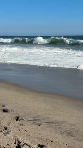 The video shows a sandy beach with footprints visible in the foreground. Gentle waves are breaking on the shore, with white foam spreading across the wet sand. The ocean water is a deep blue, transitioning to green closer to the shore, and the sky above is clear and bright blue.