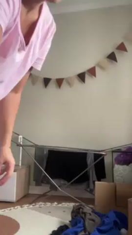 In a medium shot, a woman is standing in a room, showcasing her pajamas. She is wearing a light pink short-sleeved t-shirt and white pants with black polka dots. She is also wearing white socks. The woman has her hair pulled back in a ponytail. She is demonstrating the fit of her t-shirt by holding the sides out and giving a small twirl. The background of the room includes a clothes drying rack with clothes hanging on it, cardboard boxes, a small cabinet, a rug, and a wall decorated with triangular flags of various colors.