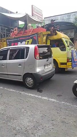 A yellow truck with a large sound system mounted on its back is parked on a street. People are sitting on top of the sound system. A silver car is parked behind the truck. A scooter is in the foreground, with a person riding it. The street is paved with gravel. The sky is overcast.
