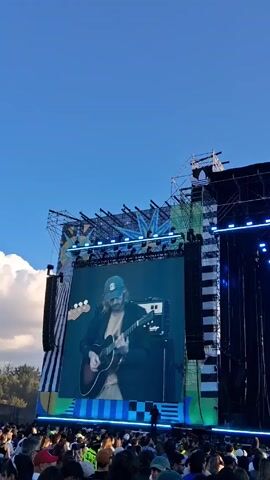 The video shows a large crowd of people at an outdoor concert under a clear blue sky. A large stage is visible, featuring a screen displaying a performer playing an acoustic guitar. The screen also shows the words "UN MUNDO DISTINTO". Lighting rigs and speakers flank the stage. Behind the crowd, trees and a Ferris wheel can be seen in the distance. The crowd is densely packed, and people are wearing a variety of casual clothing, including hats and sunglasses.