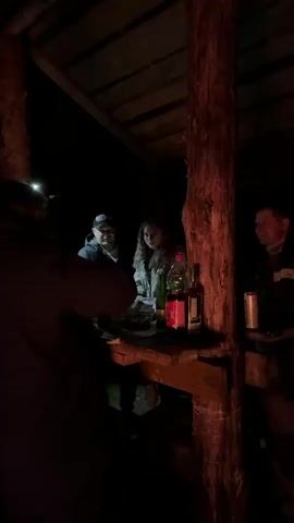 The video shows a group of four people standing around a wooden table inside a dark, rustic, open-sided structure at night. The structure has a crude wooden roof and is supported by thick wooden posts. The table is cluttered with bottles, cans, and food. One person is wearing a headlamp. A man in a black jacket and baseball cap sits at the table. A woman stands beside him, and another man stands to the right side of the table. One person stands with their back to the camera on the left. The scene is dimly lit.