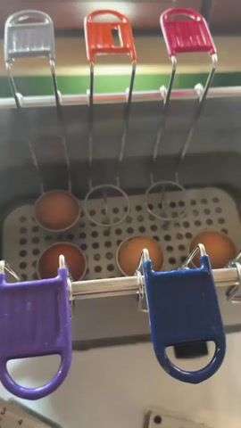 The video shows a close-up of a device for cooking eggs, likely a commercial egg cooker, with several eggs in individual metal holders suspended over a heating element. The egg holders have colored plastic handles, one purple, one blue, one silver, one orange, and one red. The device is set in a stainless steel container with a perforated bottom. In the background, a person's hand is seen interacting with a small hourglass timer with three separate bulbs, labeled with minutes and cooking levels: MIN, SOFT, MEDIUM, and HARD. The timer bulbs are filled with sand, with one bulb nearly empty, another halfway full, and the third almost full. The device appears to be on a white tablecloth, and a piece of black electronic equipment with the brand name "ggm gastro" is visible. Two small rectangular tags, possibly instructions or labels, are on the tablecloth near the timer.