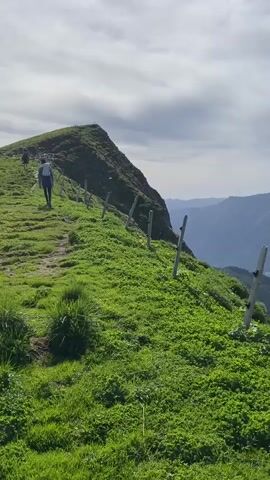 A person walks along a grassy mountain path with a fence made of wooden posts on the right. The path leads up to the left and then curves out of view. The background shows a vast expanse of green mountains and a turquoise lake under a cloudy sky. The scene is a scenic view of a mountain landscape.