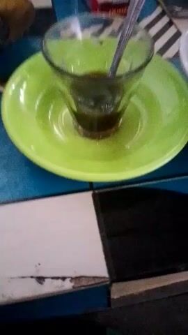 A close-up shot shows a glass of dark liquid, likely coffee, sitting on a bright green saucer. A spoon rests inside the glass, its handle slightly visible above the rim. The saucer and glass are placed on a table with a checkered surface composed of blue, white, and black tiles. Some of the white tiles appear worn and damaged at the edges. The lighting suggests an indoor environment, possibly a cafe or a home. No people are visible in the frame, and no specific actions are taking place beyond the static arrangement of the objects.