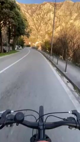 A first-person perspective video shows someone riding a bicycle on an asphalt road. The road curves gently uphill ahead. A white line marks the edge of the road on the left. On the right side of the road, there's a narrow sidewalk separated from the road by short, thin, vertical poles. Sparse vegetation and trees are present on both sides of the road. In the background, a large, rocky mountain dominates the scene, illuminated by sunlight. The bicycle's handlebars and front tire are visible in the lower part of the frame. The sky is clear and blue.