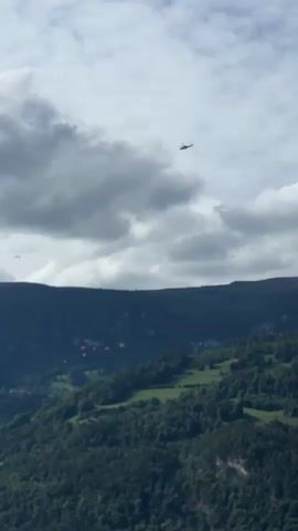 The video shows a wide aerial view of a mountainous landscape under a cloudy sky. In the distance, a small airplane is visible flying in the sky. Below the airplane, several paragliders are descending, appearing as small red dots against the green forested mountainside. A few scattered buildings and houses are visible nestled within the slopes, and the overall impression is one of a serene, natural environment where recreational activities are taking place.