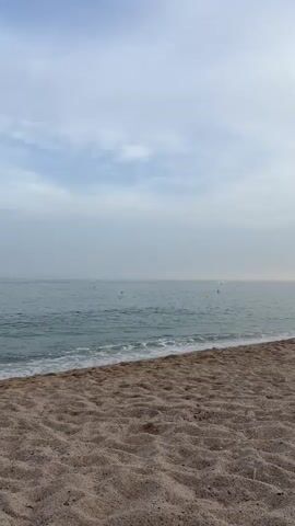 The video shows a serene beach scene. The sand in the foreground is light brown and textured with small pebbles and shells. The ocean is a calm turquoise, with gentle waves lapping at the shore. Yellow buoys are scattered across the water, marking a swimming area. The sky is overcast with soft gray clouds, creating a diffused light. The horizon is a clear, straight line separating the sea and sky. There are no people or animals visible in the frame. The overall atmosphere is peaceful and tranquil.