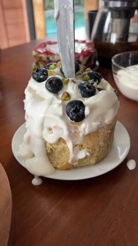 A close-up shot shows a single-serving dessert sitting on a small white plate on a wooden table. The dessert is a small cake topped with white frosting, blueberries, and small green nuts. A large knife is impaled in the center of the cake. In the background, another dessert and a glass bowl filled with frosting are partially visible. A glass coffee press stands behind the bowl of frosting. The person filming appears to remove the knife and laughs.
