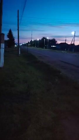 The video shows a rural road at dusk. There are streetlights illuminating the road, and the sky is a deep blue with hints of pink and orange from the setting sun. Grass and trees line the side of the road, and in the distance, there are houses. The camera is handheld and appears to be moving, as the image is slightly blurred. The audio consists of ambient sounds and what sounds like people talking in Russian.