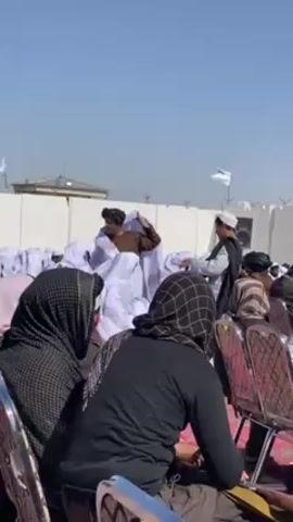 The video shows an outdoor scene with a group of men in traditional clothing. Some men are wrapping white cloth around what appear to be seated chairs. Some audience members are sitting and watching. The environment suggests a formal gathering, with a clear sky and flags in the background. The scene unfolds on a paved ground with a partial building in the background.