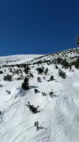 The video shows a chairlift ascending a snowy mountain. The sky is clear and blue. Other skiers can be seen on the slope below, interspersed with small trees.