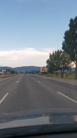 The video shows a first-person view from inside a car driving on a highway. The road has multiple lanes and is bordered by grassy fields and trees. In the distance, mountains are visible under a clear sky with some clouds. On the right side of the road, there is a large billboard featuring a woman wearing sunglasses. Further down the road on the left, there are signs and some industrial structures. The lighting suggests it is either early morning or late afternoon.