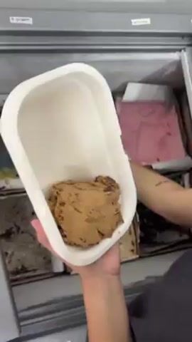 A person is using a small, silver scoop to transfer ice cream from rectangular containers in a freezer into a white container. The freezer contains several different flavors of ice cream, including chocolate, berry, and what appears to be salted caramel. The person has a tattoo on their left arm. The freezer is white and grey.