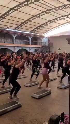 A group of women are engaged in a step aerobics class. They are all wearing athletic attire and are positioned on individual step platforms. The class is taking place indoors under a covered structure with a translucent roof, and a multi-level building is visible in the background. The participants are performing synchronized movements, raising their arms and stepping up and down on the platforms. The overall atmosphere appears energetic and focused.