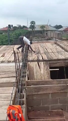The video shows ongoing construction on a building, possibly a house, in a suburban or residential area. There are several construction workers on the roof, which is covered with wooden planks and reinforced with steel rebar. Some workers are bending and tying rebar, while others are working with the wooden planks. The surrounding area consists of other houses and buildings, as well as some vegetation and a cloudy sky.