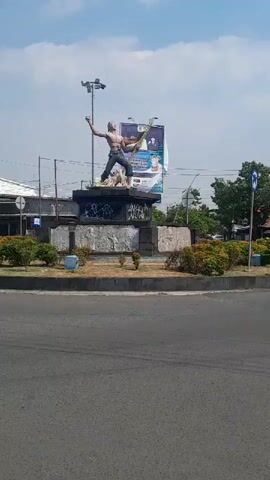 A video shows a roundabout with a statue in the center. The statue depicts a muscular figure, seemingly male, with arms raised. He appears to be in motion, potentially as part of a dynamic pose. The statue is on a stone pedestal, which is surrounded by bushes. Behind the statue, a large billboard is visible, featuring an image and text, though the details are unclear. The roundabout itself is paved, with visible road markings. The surrounding environment includes buildings, possibly shops or commercial establishments, and some trees. The sky is visible, suggesting it is daytime. There are no people or vehicles in close proximity to the statue or its immediate surroundings.
