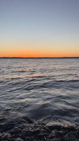 The video shows a calm body of water with gentle waves lapping at the shore. The sky above transitions from a clear pale blue at the top to a vibrant orange horizon where the sun is setting. The water reflects the colors of the sky, creating a serene and peaceful atmosphere. The shore in the foreground appears to be rocky or pebbly.