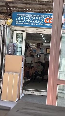 This media file shows the storefront of an electronics store called "ТехноСон" (TechnoSon) in Kryzhanivka, Ukraine. The store's sign above the entrance reads "ИНТЕРНЕТ МАГАЗИН БЫТОВОЙ ТЕХНИКИ" (Internet store of household appliances) and "Минимальные цены. Максимальный сервис." (Minimal prices. Maximum service.).

The scene is set outdoors, likely during the daytime, with natural light illuminating the area. The store appears to be a small, independent business. Inside, a person is seated in an office chair, seemingly engaged with a device they are holding, possibly a phone or tablet. They are wearing a t-shirt and shorts, suggesting a casual atmosphere and warm weather.

The storefront is partially open, revealing shelves and items displayed inside the shop. There are also several large cardboard boxes stacked outside the entrance, indicating recent deliveries or inventory. The overall impression is that of a typical small retail establishment, possibly in a market or a less bustling commercial area. The details suggest a normal business day.