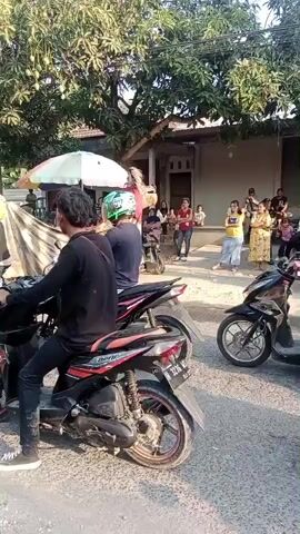 A group of people are gathered outdoors, possibly for a parade or celebration. Several motorcycles are visible, some with riders. In the center of the frame, two figures appear to be part of a traditional costume, possibly a "kuda lumping" or similar performance, covered in a light-colored cloth. One figure, on the right, is partially visible and seems to be wearing a mask or headdress. There are spectators in the background watching the event. The environment appears to be a street lined with trees and houses.