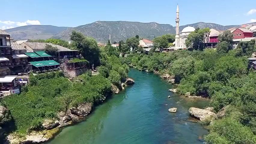This media file captures a scenic view of the Neretva River flowing through a vibrant green landscape in Mostar, Bosnia and Herzegovina. The river, with its clear turquoise waters, is flanked by lush vegetation and rocky outcrops. On the left bank, several restaurants with green awnings are nestled into the hillside, suggesting a lively atmosphere. People can be seen enjoying the outdoors at these establishments.

Dominating the right bank of the river is a prominent mosque with a tall, slender minaret, its white structure standing out against the blue sky. Adjacent to the mosque are traditional buildings with red roofs, typical of the region's architecture. In the background, a range of imposing mountains stretches across the horizon, adding to the dramatic and picturesque scenery.

The time of day appears to be mid-morning or early afternoon, given the bright sunlight casting clear shadows and the vibrant colors. The weather seems fair and warm, ideal for outdoor activities. The overall impression is one of natural beauty combined with historical and cultural landmarks. There is no visible text within the frame.