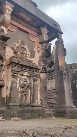 The video shows the exterior of a large, intricately carved ancient stone structure, likely a temple or monument. The architecture features massive stone pillars, detailed carvings of figures and geometric patterns, and multiple levels of doorways and alcoves. In the foreground, on the ground, are a pair of sandals, suggesting recent human presence. The environment appears to be an archaeological site with rough, rocky terrain. In the background, several people are visible, some walking around and observing the structure, while others are gathered in groups. The sky is overcast with clouds. The camera pans slowly to reveal more of the structure and its surroundings.