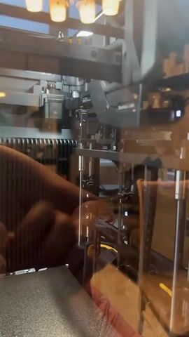 This is a close-up, interior shot of a 3D printer. The printer is a "Bambu Lab" model, as indicated by the text on a grey component. The printer has a large print bed in the foreground and various mechanical parts, including gears, belts, and an extruder assembly, visible above it. The lighting appears to be artificial, suggesting an indoor setting. The focus is on the intricate workings of the printer, with some reflections visible on a clear enclosure, obscuring the full view of the surrounding environment. There are no people visible in the frame. The activity depicted is the internal machinery of a 3D printer, implying it is either in operation or ready for use. The time of day and weather are not discernible. The location is Pervouralsk, Russia, but there are no specific visual cues in the image to confirm this. The overall impression is one of technical complexity and precision.
