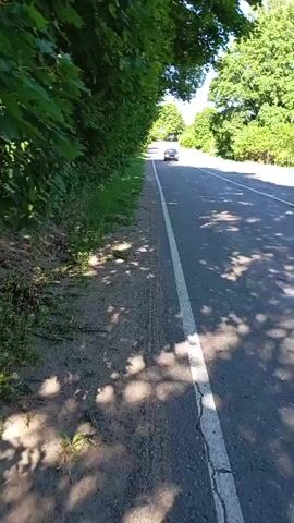 This video depicts a view from a roadside in Retyun, Russia, likely during daytime with clear weather given the bright sunlight and shadows. The scene is dominated by lush green trees lining the left side of the road, casting dappled shadows onto the dirt shoulder. The road itself is paved with a white line marking the lane.

The primary activity is vehicular movement. A dark-colored car is seen driving away from the camera down the road. The car appears to be the sole object of focus on the road itself, making its presence notable. The setting is rural or semi-rural, characterized by the abundant greenery and the open road.

There is no visible text in this short video clip. The overall mood is peaceful and ordinary, capturing a common roadside scene.