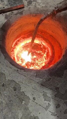 The video shows a close-up, top-down view of a circular, dark-colored industrial furnace or crucible. The interior is filled with intensely bright, glowing molten material, primarily orange and white, indicating extreme heat. The outer edge of the container is a duller reddish-orange, suggesting high temperatures radiate outwards.

A metal pipe or rod is visible extending into the molten material from the top-right. As the pipe is immersed, it causes a violent reaction within the melt, generating a brilliant white flare of light and significant amounts of smoke or vapor. The molten material appears turbulent and agitated where the pipe interacts with it. The exterior of the furnace is a cracked, grayish surface, consistent with wear and tear from industrial use. The overall scene depicts a high-temperature industrial process, likely involving melting, refining, or treating metals. No people or visible text are present in the frame. The event takes place in what appears to be an industrial setting.