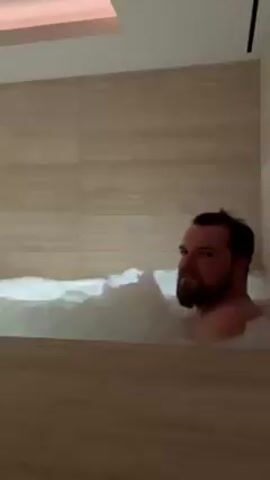 A bearded man is sitting in a bubble bath. The bathroom walls are covered in a light colored stone pattern. An overhead light fixture illuminates the room. The man looks directly at the camera. He appears relaxed. The bath is filled with dense, white bubbles.