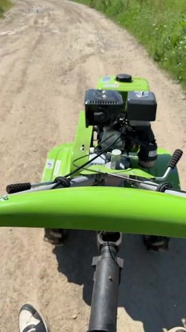A first-person perspective shot shows a person driving a green two-wheel tractor along a dirt and gravel path. The tractor's engine is visible in the foreground, with the handles extending towards the camera.  The path is lined with green vegetation, including some red poppy-like flowers, on both sides. In the distance, a building and trees are visible, suggesting a rural or agricultural setting. The video is taken during the day, with clear sunlight. The person driving the tractor is wearing a white sneaker that is partially visible. The tractor moves steadily along the path.